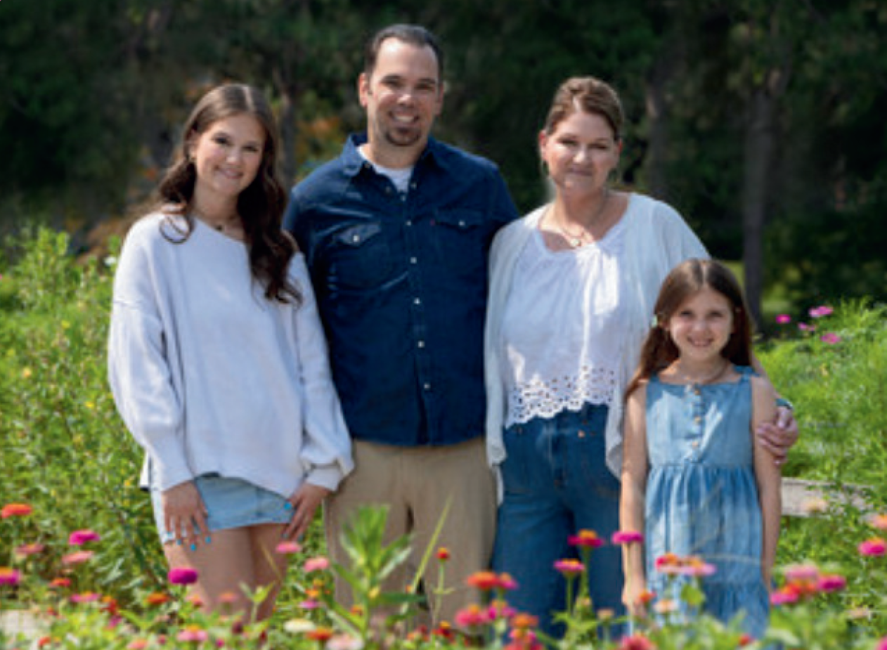A family of four posing together outdoors in a garden with colorful flowers and green foliage in the background.